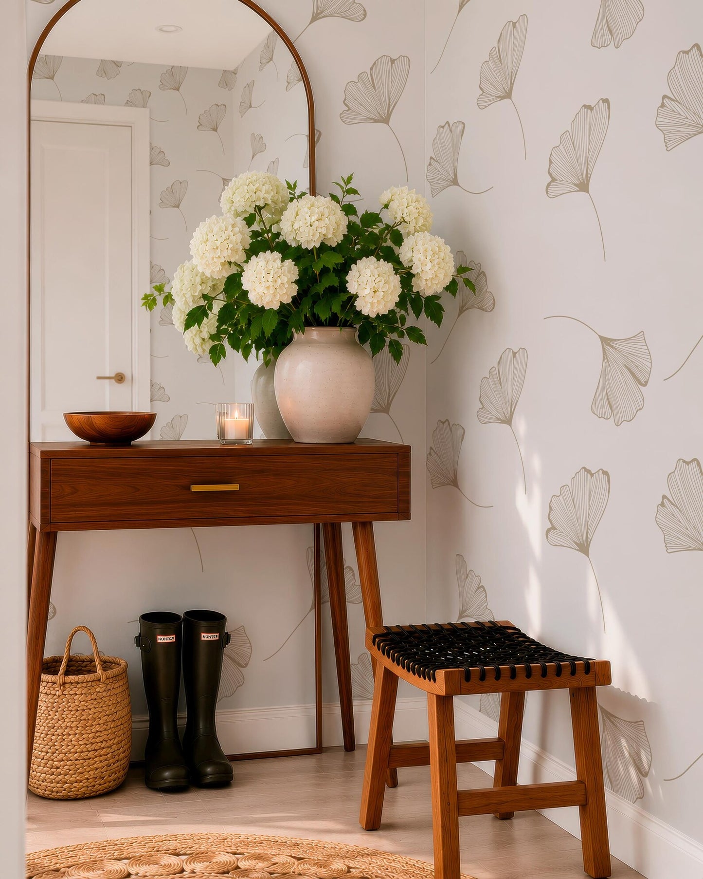 a cozy corner of a room with a wooden table, a vase of white hydrangeas, a pair of black rain boots, and a woven basket. The table has a drawer and a mirror above it, and the wall behind the table has a repeating pattern of white flowers.