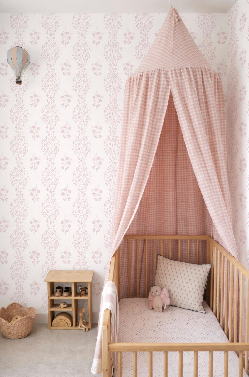 A cozy nursery with a wooden crib, a canopy bed, and a small wooden table. The walls are covered in a pink and white floral wallpaper, and there is a basket on the floor.