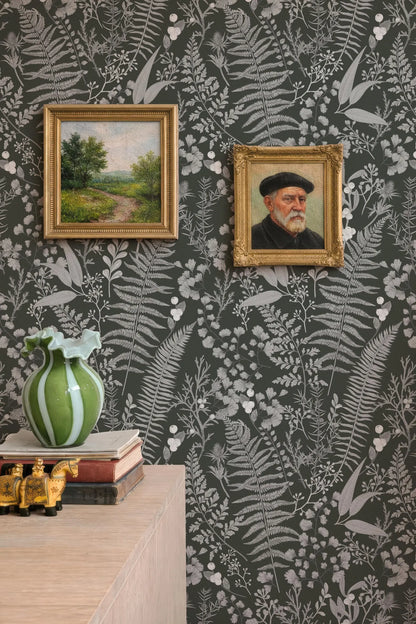 a wall with two framed paintings hanging on it, one depicting a landscape and the other a portrait of a man. There is also a green vase and a stack of books on a wooden surface below the paintings.