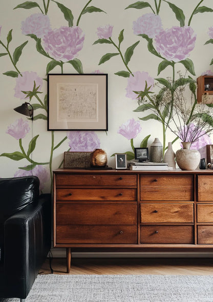 a living room with a wooden dresser, a black leather couch, and a framed floral wallpaper. The dresser has a wooden top and multiple drawers. The wall behind the dresser is decorated with a floral wallpaper, and there is a framed picture hanging on the wall. The room also contains a vase with flowers and some books on the dresser.